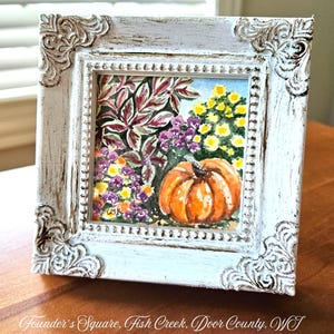 May include: A square watercolor painting of an orange pumpkin surrounded by purple and yellow flowers and dark foliage, housed in a white distressed ornate frame with corner scrollwork. The text Founder's Square, Fish Creek, Door County, WI appears.
