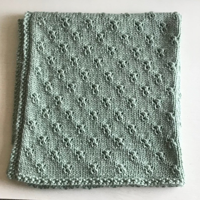 May include: A soft, sage green knitted baby blanket with a diamond pattern.