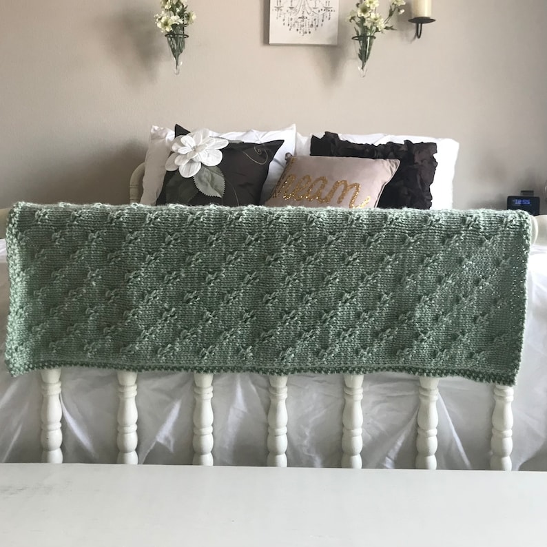 May include: A sage green knitted throw blanket with a diamond pattern draped over a white bed frame.