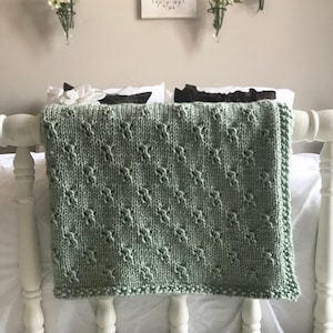 May include: A sage green knitted throw blanket with a diamond pattern, draped over a white bed frame.