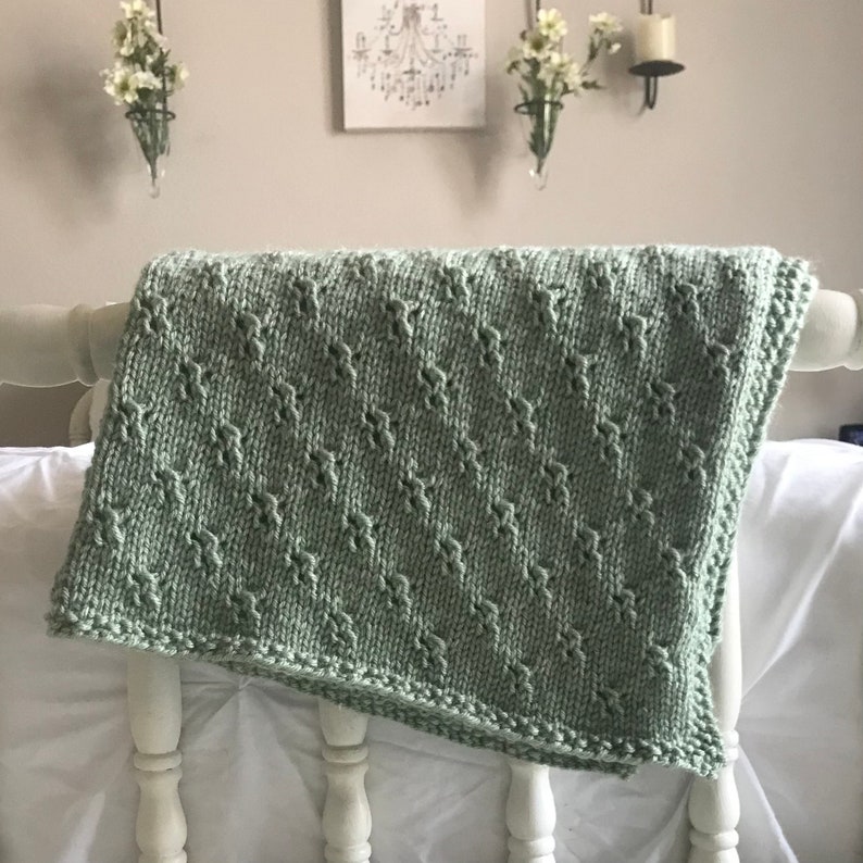 May include: A sage green knitted baby blanket with a delicate diamond pattern. The blanket is draped over a white wooden chair.