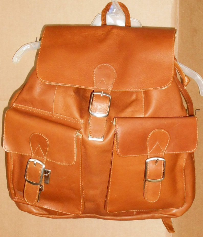 Pu&ograve; includere: Uno zaino in pelle marrone con due grandi tasche anteriori e due fibbie. Lo zaino ha una patta con chiusura a fibbia.