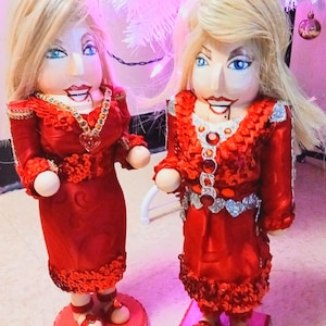 May include: Two wooden nutcracker dolls dressed in red sequined dresses with white accents. The dolls have blonde hair and painted faces. The dolls are standing on red and pink bases.