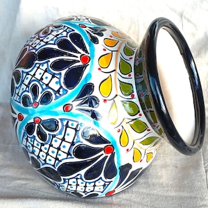 Puede incluir: Un jarrón de cerámica redondo con base blanca y patrones pintados a mano en azul oscuro, turquesa, amarillo, verde y rojo. El diseño incluye motivos florales, patrones de cuadrícula y formas de hojas. El jarrón tiene un borde negro brillante.