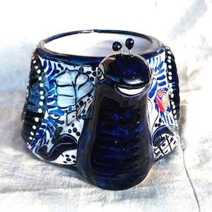May include: A ceramic planter shaped like a snail with a glossy dark blue head and antennae. The shell features intricate hand-painted patterns in shades of blue, white, and black, including floral motifs and geometric lines on a white background.
