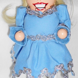 May include: A wooden doll with blonde hair wearing a blue dress with silver trim and standing on a blue base with silver trim.