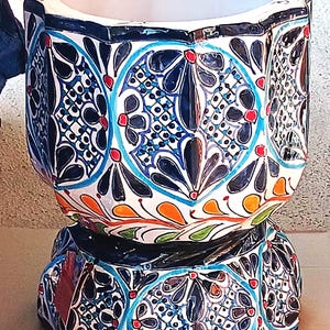 May include: A ceramic planter featuring a scalloped rim and a wide, cylindrical body that tapers toward a pedestal base. The exterior is decorated with intricate hand-painted patterns in dark blue, light blue, orange, and green on a white background.