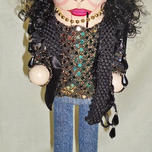 Puede incluir: Un cascanueces decorativo con una cara pintada, cabello negro rizado y un atuendo negro con lentejuelas. La muñeca lleva vaqueros azules y zapatos azules, de pie sobre una base negra. Un collar dorado está alrededor del cuello.