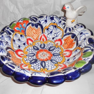 Puede incluir: Un bebedero para pájaros de cerámica azul y blanco con un pájaro blanco posado en el borde. El bebedero está decorado con diseños florales coloridos y tiene un borde festoneado.