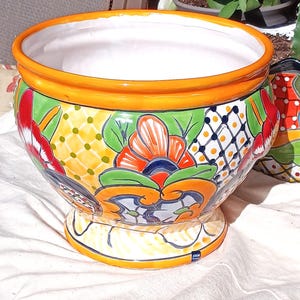May include: A round ceramic planter with a wide orange rim and a white interior. The exterior features a vibrant hand-painted pattern of red, orange, and green floral motifs, yellow dotted sections, and blue grid designs on a curved body.