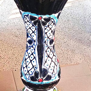 May include: A ceramic pedestal stand featuring a flared, scalloped rim and a tapered body. The surface is decorated with hand-painted black, blue, and white lattice patterns, accented by red dots, and a base adorned with green and orange leaf motifs.