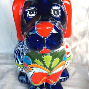 May include: A ceramic dog figurine featuring a glossy finish with a dark blue base color. The dog has bright red ears and nose, with hand-painted floral and geometric patterns in green, orange, and white across its body and chest area.