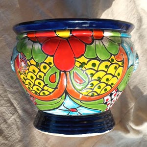 May include: A ceramic planter featuring a vibrant hand-painted floral pattern in red, yellow, green, and light blue. The bowl-shaped vessel has a dark blue rim and a matching dark blue pedestal base, with small side handles integrated into the design.