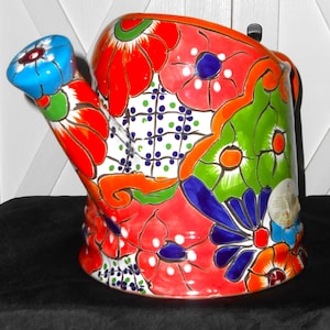 May include: A colorful ceramic watering can with a floral design. The can is painted in shades of red, orange, yellow, green, blue, and white. The spout is curved and the handle is shaped like a flower.