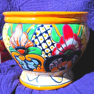 May include: A round ceramic planter featuring a vibrant hand-painted design with bright orange, green, red, and yellow floral motifs, a black grid pattern, and a solid orange rim. The base is flared, and a small product tag is attached to the side.