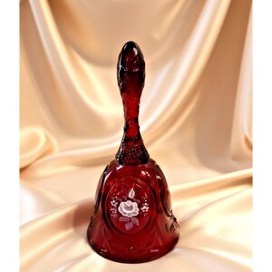 Puede incluir: Una campana de cristal de color rojo rubí intenso con un mango fino y texturizado y una base acampanada. El exterior de la campana está decorado con un motivo floral blanco pintado a mano que muestra una rosa central con hojas.