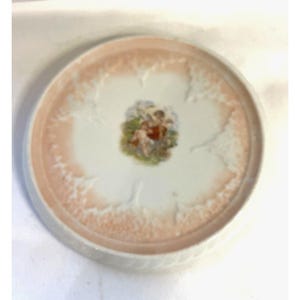 Puede incluir: Un plato redondo de cerámica blanca con un diseño floral rosa y blanco alrededor del borde. El centro del plato presenta una pequeña ilustración colorida de querubines. El plato tiene un borde festoneado.