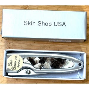 Può includere: Un coltello color argento con un manico con motivo a pelle di serpente, presentato in una scatola bianca. La scatola è etichettata "Skin Shop USA". Il coltello è in una scatola bianca.