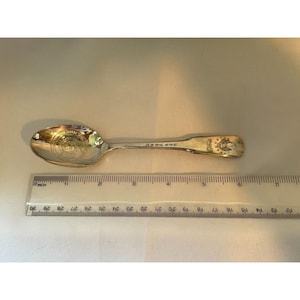 May include: Silver teaspoon with a gold-plated design featuring a crest with the words "ERE HONI" on the handle. The spoon is 6 inches long.