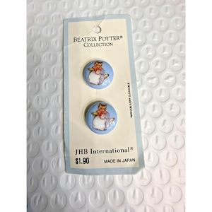 Op de afbeelding: Twee ronde knopen uit de Beatrix Potter Collection. Elke knoop heeft een cartoonkat op een lichtblauwe achtergrond. De kaart leest "JHB International" en "MADE IN JAPAN".