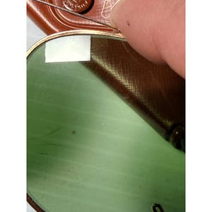 May include: A pair of sunglasses with a gold-toned metal frame and light green tinted lenses. A small rectangular white sticker is attached to the upper left corner of the lens. The sunglasses are partially resting against a brown textured case.