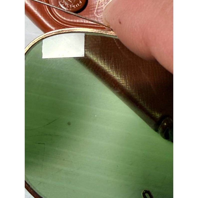May include: A pair of sunglasses with a gold-toned metal frame and light green tinted lenses. A small rectangular white sticker is attached to the upper left corner of the lens. The sunglasses are partially resting against a brown textured case.