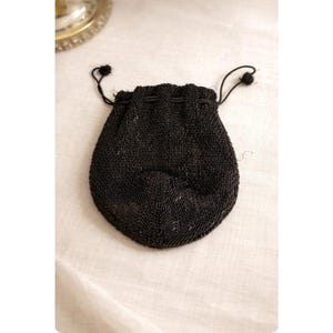 May include: A small, black beaded drawstring bag. The bag is oval-shaped with a gathered top and a black cord drawstring. The beads appear to be tightly sewn together, creating a textured surface. The bag is likely vintage or antique.