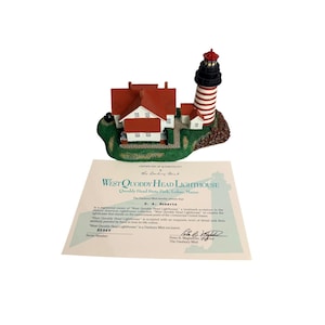 Pode incluir: Uma réplica em miniatura do Farol de West Quoddy Head, com uma torre listrada em vermelho e branco, uma casa com telhado vermelho e um certificado de autenticidade. O farol está sobre uma base verde.