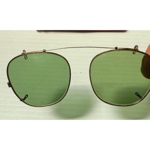 May include: A pair of clip-on sunglasses featuring two rounded, light green tinted lenses held together by a thin, gold-toned metal bridge. The frame includes small metal clips at the top and sides for attachment to existing eyewear frames.