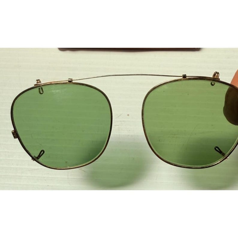 May include: A pair of clip-on sunglasses featuring two rounded, light green tinted lenses held together by a thin, gold-toned metal bridge. The frame includes small metal clips at the top and sides for attachment to existing eyewear frames.