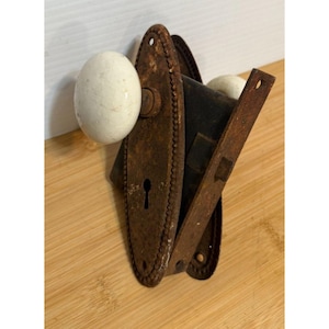 Puede incluir: Juego de herrajes de puerta antiguos con una placa trasera oxidada de forma ovalada con borde de cuentas. Incluye un pomo de cerámica blanca y una cerradura a juego. El juego es de metal y muestra signos de edad y desgaste.