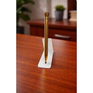 Könnte beinhalten: Ein goldfarbener Stift mit detailliertem Design, der in einem weißen Halter steht. Der Stift hat eine Spitze und eine dekorative Oberseite. Der Stift wird auf einem Holzschreibtisch ausgestellt.