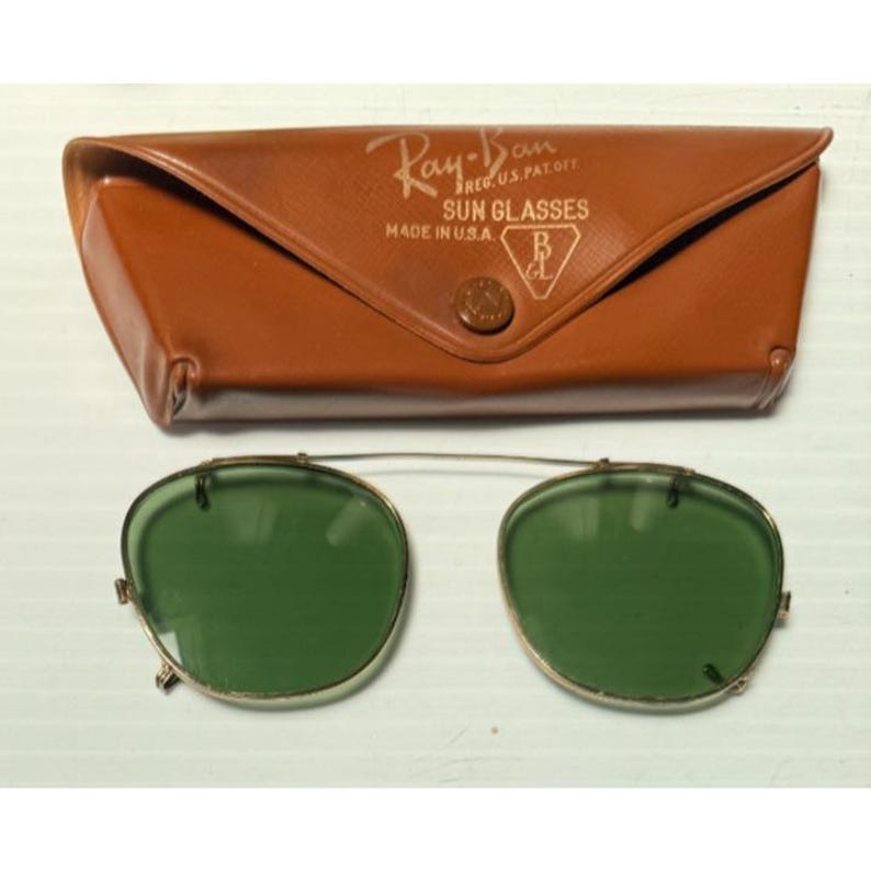 May include: A pair of clip-on sunglasses with green lenses and a thin gold-tone metal frame, accompanied by a brown leather-like case. The case features the text Ray-Ban REG.U.S.PAT.OFF. SUN GLASSES MADE IN U.S.A. and a B&L logo on the front flap.