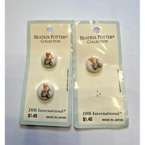 Puede incluir: Tres botones redondos blancos con una ilustración impresa de Peter Rabbit, montados en dos tarjetas de presentación de cartón azul claro y crema etiquetadas como Beatrix Potter Collection y JHB International, con texto que indica Made in Japan.