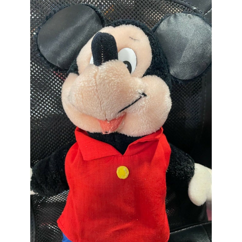Pode incluir: Um brinquedo de pel&uacute;cia do Mickey Mouse vestindo uma camisa vermelha com um bot&atilde;o amarelo. O brinquedo tem orelhas pretas, um nariz preto e um rosto rosa.