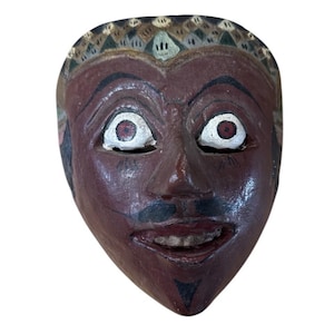 May include: A carved wooden mask with a dark reddish-brown face, white eyes with red pupils, and a black moustache. The mask has a smiling mouth and a decorative patterned headdress. The mask is likely a cultural or theatrical item.