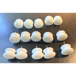 May include: A collection of fourteen white ceramic cabinet knobs arranged in three rows. Each knob features a smooth, rounded, mushroom-shaped head and a metal screw protruding from the back for installation, providing a uniform look for furniture.