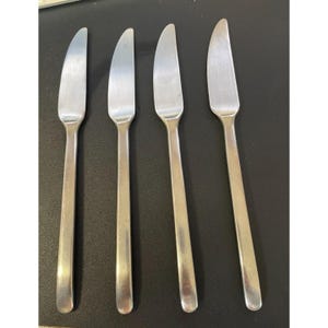 May include: Four stainless steel butter knives with curved blades and long, slender handles. The knives are arranged in a row, reflecting light, and are set against a dark background. Each knife measures approximately 8 inches in length.