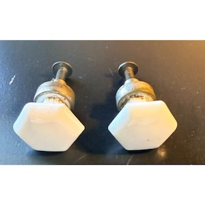 May include: Two hexagonal white ceramic cabinet knobs with metallic bases and threaded screws. Each knob features a smooth, glossy white top surface in a six-sided shape, connected to a circular metal mounting piece with a visible screw stem.