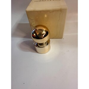 May include: A gold-colored perfume bottle stopper with a spherical top and a cylindrical base. The stopper is positioned in front of a beige box with the brand name "Courreges" printed on it.