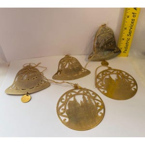 Può includere: Una collezione di ornamenti natalizi in metallo color oro. Gli ornamenti includono forme a campana e disegni circolari con motivi traforati. Un ornamento riporta l'anno "1981". Gli ornamenti sono infilati con spago.