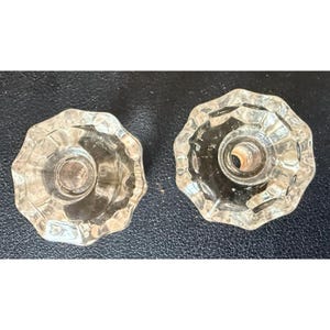 May include: Two clear glass knobs with a multi-faceted, star-shaped design. Each knob features a circular center with a small hole for mounting. The glass is transparent and reflects light, showing a distinct scalloped edge around the perimeter.
