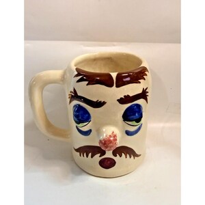 Könnte beinhalten: Eine cremefarbene Keramiktasse mit Gesichtsdesign. Die Tasse hat blaue Augen, eine rote Nase und einen braunen Schnurrbart und Augenbrauen. Der Griff ist ebenfalls cremefarben.