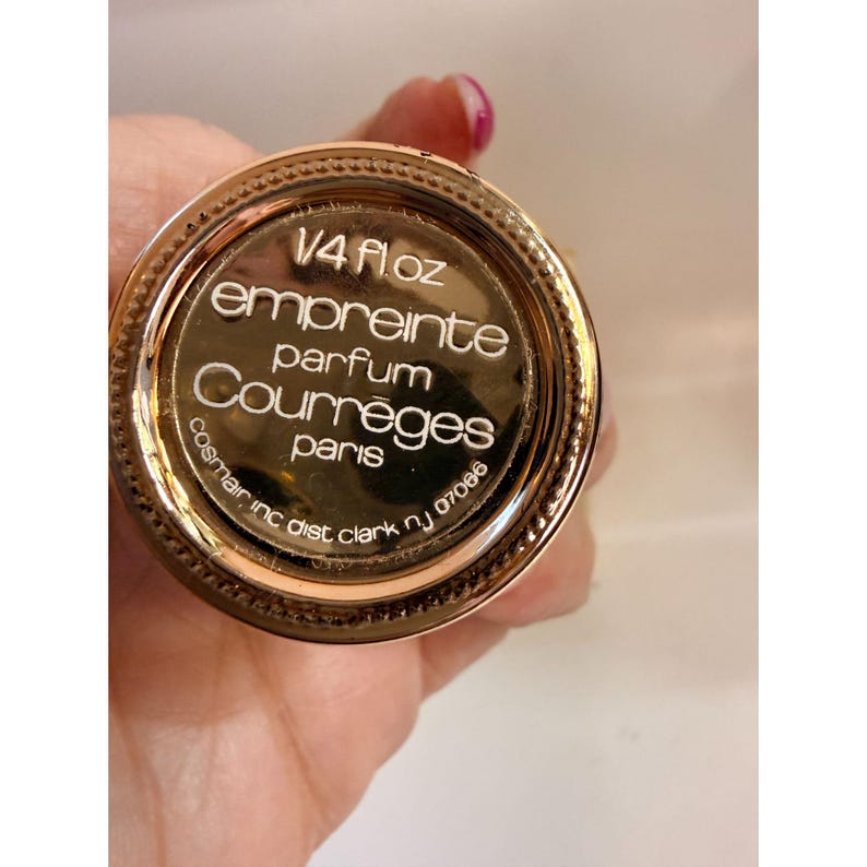 May include: A gold-colored perfume bottle base with the text "1/4 Fl Oz empreinte parfum Courr&egrave;ges paris" in white. The bottle base is circular with a textured edge and is held by a hand.