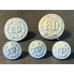 May include: A set of five silver-toned metal buttons of varying sizes, each featuring the embossed letters FD in the center surrounded by a detailed wreath design. The buttons have a circular shape with a raised border and a metallic finish.
