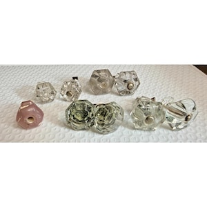 May include: A collection of nine hexagonal cabinet knobs, including eight clear glass knobs with silver-toned metal centers and one solid light pink glass knob. Each knob features a faceted design with a central hole for mounting hardware.