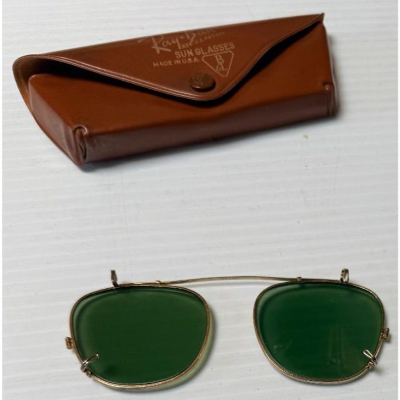 May include: A pair of gold-tone metal clip-on sunglasses with dark green lenses and a brown leather-like carrying case. The case features the embossed text Ray-Ban SUN GLASSES MADE IN U.S.A. and a B&L logo on the front flap with a snap closure.
