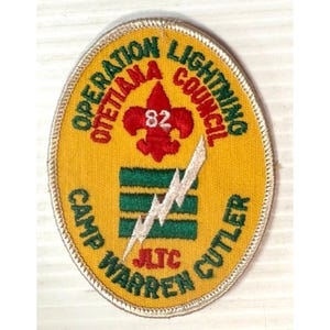 Peut inclure: Un écusson brodé de forme ovale, jaune, avec des textes et des graphiques verts et rouges. L'écusson porte les inscriptions "OPERATION LIGHTNING COUNCIL" et "CAMP WARREN CUTLER". Un fleur-de-lis rouge avec le numéro 82 est au centre, avec un éclair blanc.