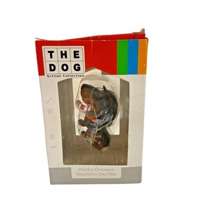 May include: A holiday ornament featuring a dachshund dog with a candy cane. The dog is black and brown with a gold string for hanging. The ornament is in a box with the text "THE DOG Artlist Collection" and "Holiday Ornament Décorations Des Fêtes".