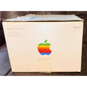 Peut inclure: Une boîte en carton blanc pour un Apple 5.25 Drive. La face avant présente le logo Apple classique aux couleurs de l'arc-en-ciel, le texte Apple 5.25 Drive en plusieurs langues et des icônes d'expédition standard dans le coin supérieur droit.